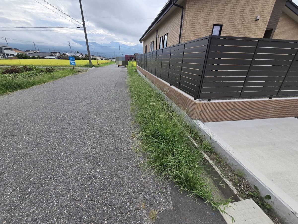 駒ヶ根市の道路縁の草刈り前:フェンス沿いに雑草が茂り縁石との境目が見えにくい状態