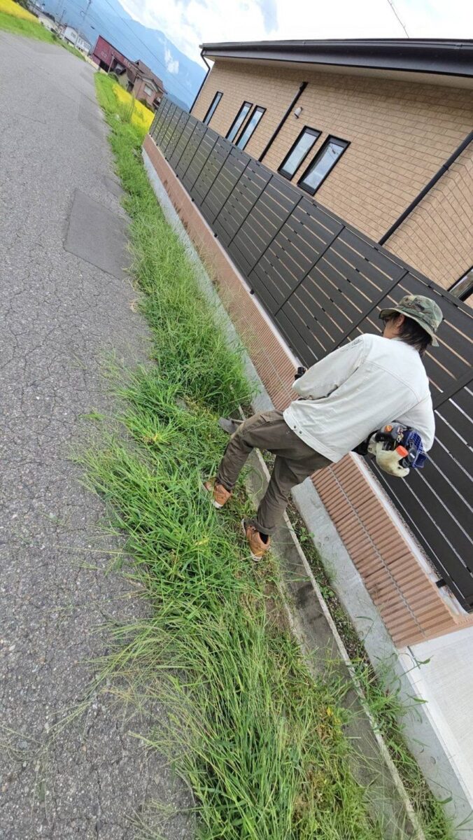 駒ヶ根市の道路縁の草刈り作業中:刈払機でフェンス際の雑草を刈り込む様子